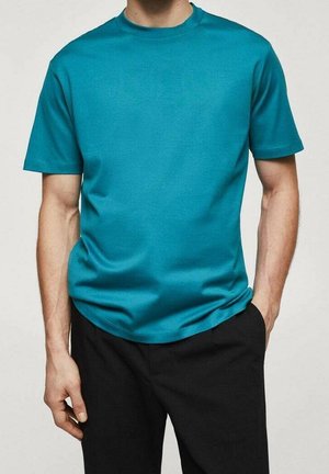 Mand iført en ensfarvet teal t-shirt med rund hals og korte ærmer samt sorte bukser, stående med den ene hånd i lommen mod en neutral baggrund.