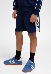 Kind draagt marineblauwe sportshorts met witte strepen aan de zijkant en blauwe sneakers met witte chevron-accenten en zwarte sokken.