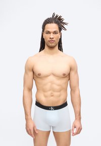 Άνδρας με κοτσίδες, χωρίς μπλούζα, που φοράει λευκά εσώρουχα Calvin Klein boxer briefs, στέκεται με το πρόσωπο προς τα εμπρός σε απλό ανοιχτό φόντο.