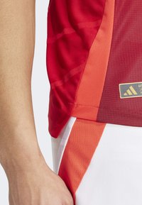 Maillot de sport rouge avec un tissu texturé, des accents orange et une étiquette de logo. Détails de la coupe des manches et de la couture visibles.