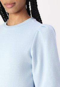 Sweatshirt azul claro com decote redondo e mangas bufantes. Textura suave e tecido liso. Design minimalista sem padrões visíveis ou acessórios.