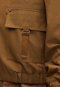 Veste marron en tissu texturé, dotée d'une grande poche avant et d'une sangle avec une boucle métallique. Détail de ceinture élastiquée.