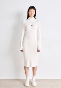 Vestido blanco de manga larga con textura acanalada, cuello alto y un pequeño parche con el logo en el pecho, combinado con zapatillas blancas y calcetines tobilleros.