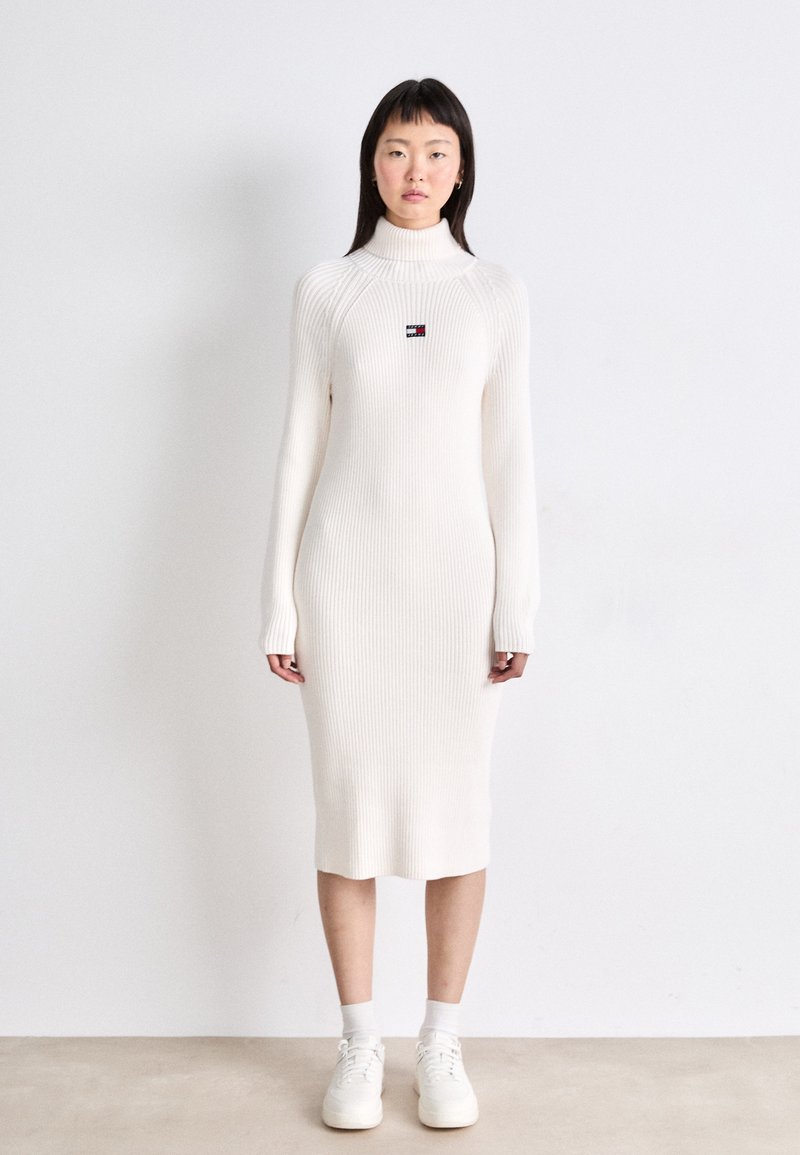 Vestido blanco de manga larga con textura acanalada, cuello alto y un pequeño parche con el logo en el pecho, combinado con zapatillas blancas y calcetines tobilleros.