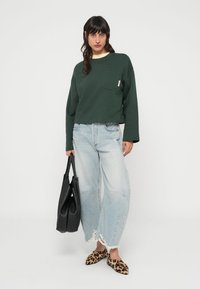 Πράσινο κοντό φούτερ με μπροστινή τσέπη, σε συνδυασμό με ανοιχτό μπλε τζιν με φαρδιά μπατζάκια και σκισίματα, και φλατ παπούτσια με λεοπάρ εκτύπωση, κρατώντας μαύρη τσάντα.