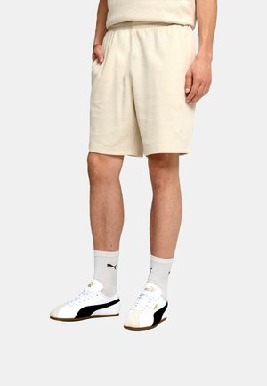 Mężczyzna w beżowych szortach, białych skarpetkach Puma i białych butach sportowych Puma z czarnymi paskami, stoi na tle jednolitego tła.