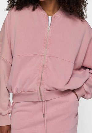 Person trägt eine rosa Zip-up-Jacke mit gerippten Bündchen und Saum, kombiniert mit passenden rosa Jogginghosen mit Kordelzug, eine Hand in der Tasche.