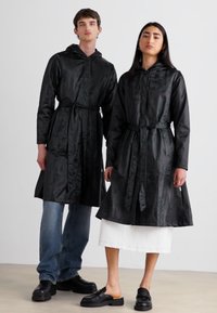 Black hooded raincoats with waist ties, featuring a satin-like texture. Styled with blue jeans and a white skirt, set against a neutral background.