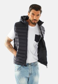Gilet con cappuccio in nave scuro con texture trapuntata, zip anteriore e tasche laterali, indossato sopra una T-shirt bianca con una tasca nera.