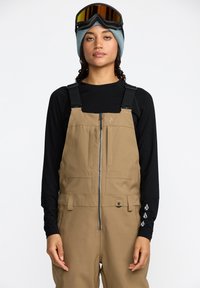 Salopette en neige beige avec des bretelles ajustables, fermeture éclair à l'avant et poches latérales, associée à un t-shirt noir à manches longues et un bonnet en tricot bleu.