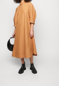Hnědress s délka po kolena, nabíranými rukávy, rovný střih a detaily s knoflíky. Nosené s černými kotníkovými botami a černou kabelkou.