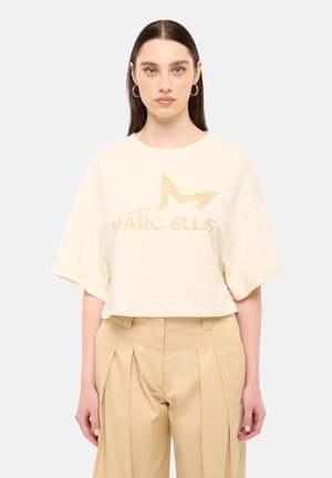 Donna con lunghi capelli scuri che indossa orecchini a cerchio dorati, una maglietta corta color crema con una stella dorata e testo, e pantaloni beige a gamba larga a vita alta.