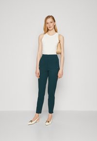 Tyrkysové nohavice na mieru so slim fit strihom, spárované s bielym topom bez rukávov. Doplnky zahŕňajú béžovú tašku a off-white lodičky s ostrou špičkou.