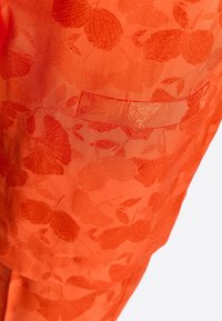 Tissu orange avec un motif floral texturé, doté d'une petite poche rectangulaire intégrée dans le design. Surface lisse.