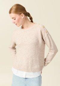 Maglione a maglia beige chiaro con collo rotondo, orlo a coste e quattro bottoni dorati decorativi sulla spalla sinistra. Materiale morbido e testurizzato.