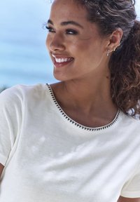 Bela bombažna srajca z okroglim izrezom, ki ima črno trikotno šivano podrobnost ob robu. Mehka tekstura in kratki rokavi.