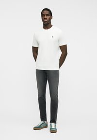 Homme debout, les mains derrière le dos, portant une chemise blanche à manches courtes, un jean gris foncé et des baskets bleu sarcelle avec des lacets blancs.