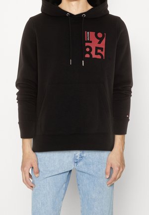 Sweat à capuche noir avec un design graphique rouge à l'avant, doté de cordons de serrage, d'une poche kangourou à l'avant et de poignets côtelés.