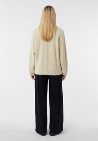 Miękki beżowy, puszysty sweter o luźnym kroju i zaokrąglonym brzegu, zestawiony z luźnymi czarnymi spodniami. Strój ma długie rękawy i swobodną sylwetkę.