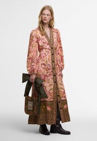 Robe longue rose à motif floral avec des manches bouffantes et des accents dorés, dotée d'une jupe brodée verte et d'un sac à main vert assorti.
