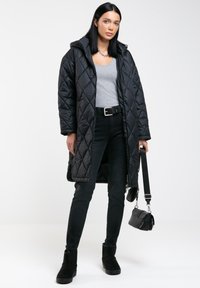 Abrigo largo acolchado negro con capucha, camiseta ajustada gris, jeans negros de tiro alto y botines negros, sosteniendo un pequeño bolso negro.