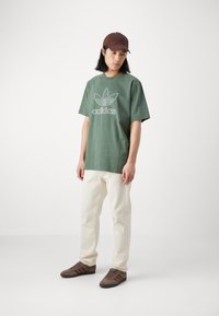 Zielona bawełniana koszulka z białym logo na piersi, luźny krój. Połączona z jasnoszarymi spodniami i brązowymi zamszowymi butami z trzema paskami.