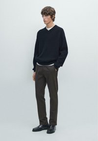 Sort, ribbet strikket sweater med V-hals, parret med mørkebrune bukser. Modellen står foran en neutral baggrund med hænderne i lommerne.