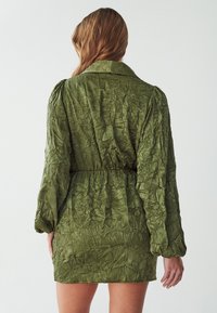 Olivegrünes, zerknittertes Kleid aus Stoff mit langen Puffärmeln, taillenbetont und kurzer Länge. Verfügt über einen spitzen Kragen und eine strukturierte Oberfläche.