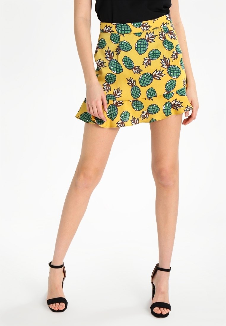 Jupe jaune à motif d'ananas, avec un ourlet volanté, fabriquée en tissu léger. Assortie à des sandales à talons hauts noires.
