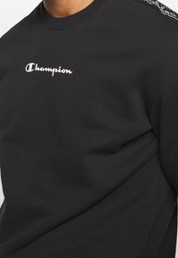 Felpa nera in cotone, con un logo bianco "Champion" sul petto e un bordo decorato lungo le cuciture delle spalle.