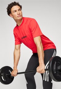 Raudona Adidas marškinė su juodu logotipu, derinami su juodomis sportinėmis kelnėmis, kurios turi baltas juosteles, keliančios štangą su svorio plokštelėmis.