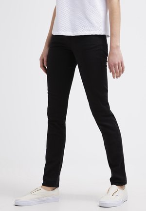 Personne portant un pantalon slim noir, un top blanc texturé sans manches et des baskets blanches à lacets, debout devant un fond uni.