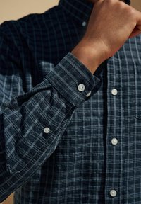 Een button-up overhemd in donkerblauw en grijs ruitpatroon, met een gestructureerde stof, oprolbare mouwen en witte knopen.