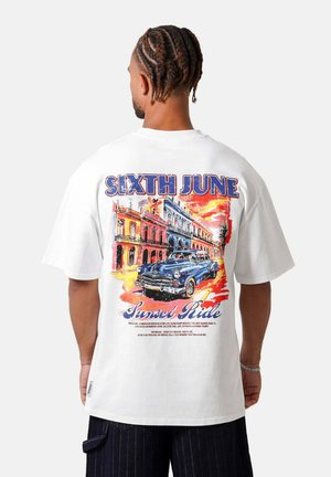 Man met gevlochten haar die een wit T-shirt draagt en een kleurrijke vintage auto en gebouwen toont met de tekst "Sixth June Sunset Ride" op de achterkant.