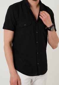 Kortärmad svart skjorta med två framfickor, bruna knappar och en mjuk textur. Bärs med vita byxor och accessoarer.