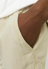 Pantalon en coton beige avec poches latérales, montrant une vue partielle d'une main reposant à l'intérieur de la poche. Tissu texturé et détails de couture soignés visibles.
