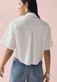 Chemise blanche à manches courtes avec un pli dans le dos et des clous décoratifs sur les manches, associée à un jean taille haute bleu clair. Accent en bijoux en argent.