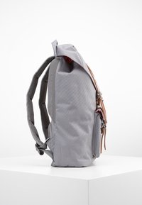 Harmaa kangaskassi, jossa on sivuvetoketjullinen tasku, ruskeat nahkaosat ja pehmustettu verkkoselkä. Muoto on pyöristetty ja siinä on säädettävät hihnat.