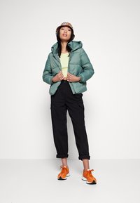 Dikke groene jas met capuchon, ritssluiting en elastische manchetten. Gepaarde met zwarte cargo-broeken en feloranje sneakers.