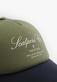 Gorra verde con visera azul marino oscuro; presenta un logotipo de texto blanco y un eslogan. El material parece tejido con un acabado texturizado. Parte trasera ajustable.