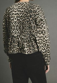 Leopardenmuster-Bluse in Beige und Schwarz, mit rüschengeschneidertem Peplumsaum, langen Ärmeln und lockerer Passform. Kombiniert mit schwarzen Hosen.