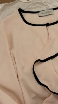 Light beige long-sleeve top with black trim along neckline and cuffs, featuring a small clasp closure at the collar and brand label "ANNA FIELD."