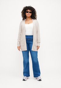 Beige knitted cardigan with open front and pocket details, paired with blue flared jeans and gray athletic shoes, on a model with curly hair.