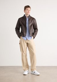 Brun skinnjakke med en tettsittende design, lyseblå stripete skjorte under, beige bukser og hvite joggesko på en nøytral bakgrunn.