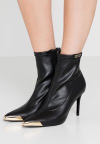 Czarne skórzane botki sięgające kostki o eleganckim wzornictwie, z ostrym noskiem i złotym metalicznym akcentem na czubku. Posiadają zamek błyskawiczny z tyłu.