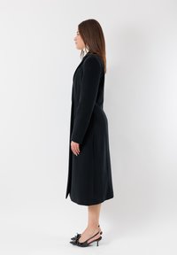 Cappotto lungo nero con un design elegante, tasche laterali e una texture liscia. Abbinato a décolleté nere con un dettaglio a fiocco.