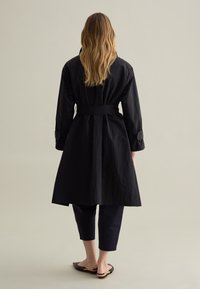 Abrigo negro de trench con cintura ceñida, mangas largas y largo hasta la mitad de la pantorrilla. Combinado con pantalones oscuros y cortos y zapatos planos. Tejido texturizado.