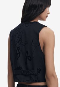 Černý bezramenní crop top s květinovou texturou, detailem knoflíku na zádech a jemným, hladkým povrchem. Vhodný pro různé příležitosti.