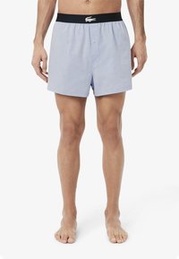 Light blue cotton boxers with a black elastic waistband featuring a white logo, single button closure, and a relaxed fit design.
