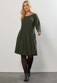 Grön långärmad klänning med rund halsringning, vid kjol och sidofickor. Bärs med svarta ankelboots, mot en neutral bakgrund.